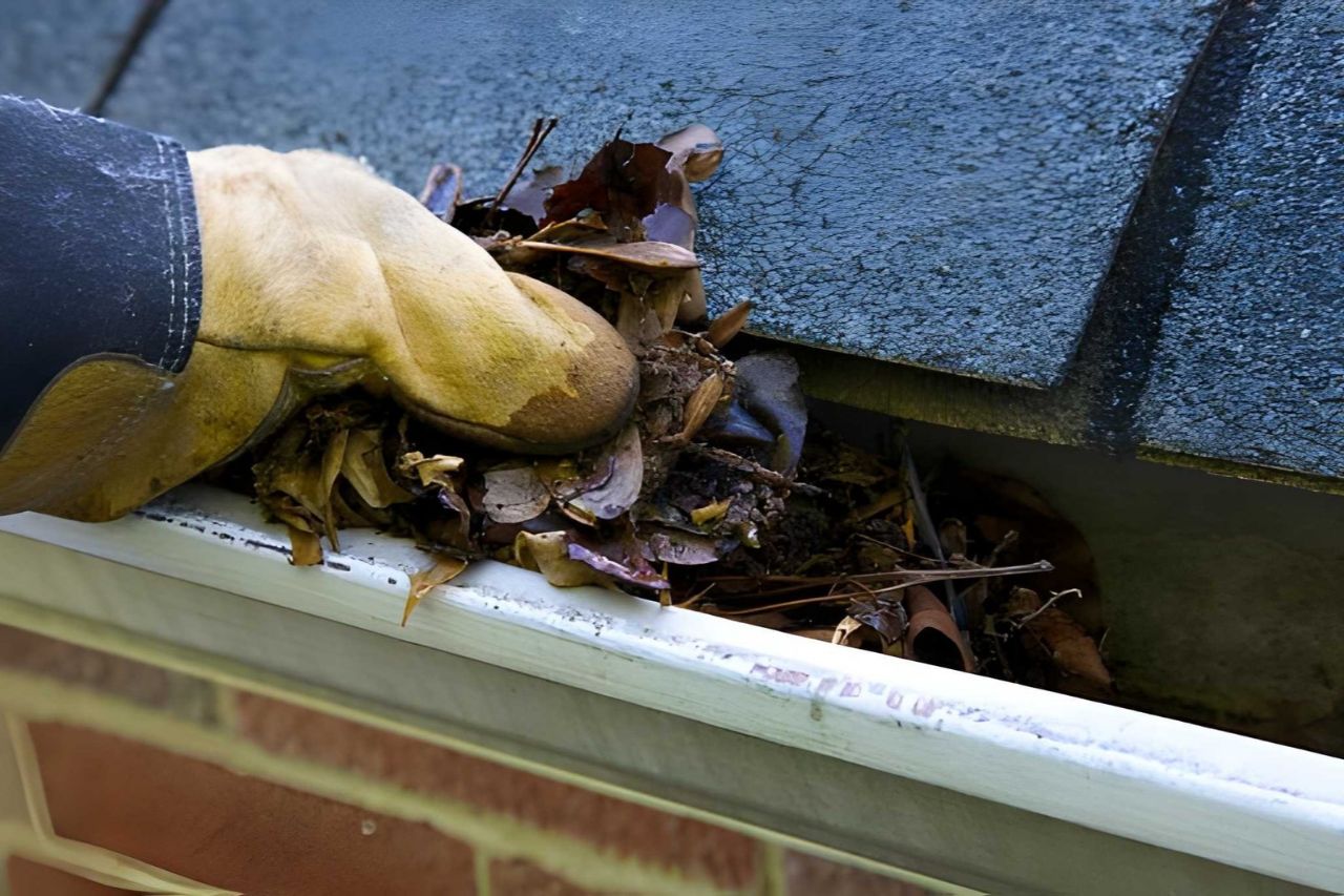 Gloved hand clearing clogged gutter with leaves and debris