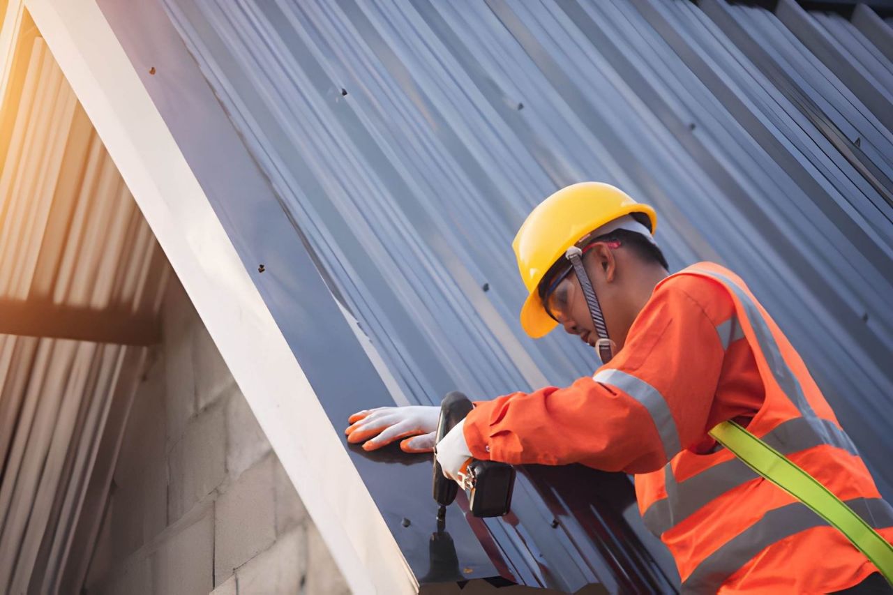 Roofer in safety gear installing metal roofing panels with a drill.