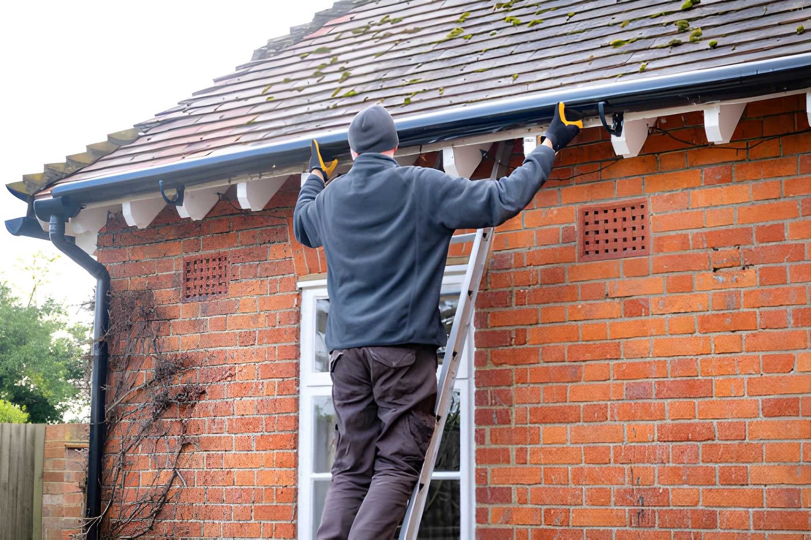 Man on ladder cleaning gutters on a brick house.
